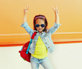 Portrait of little girl child wearing a backpack, sunglasses, jeans jacket on city street over orange background
