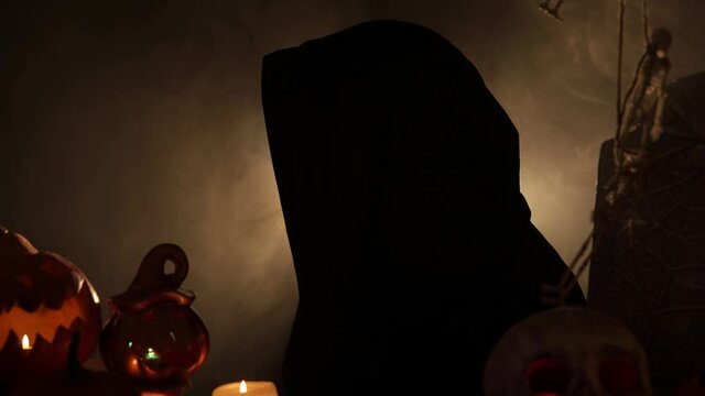 Woman in mantle with sugar skull makeup turns her head to the camera. Portrait of the girl with scary makeup among halloween decorations. Theme of halloween and santa muerte together.