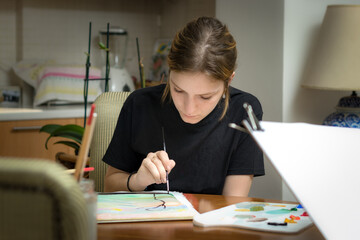 Naklejka na ściany i meble A teenage woman painting on paper with acrylic holding a painbrush sitting at home.