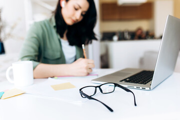 Desktop. Glasses. In the background, in defocus, a young student girl writes information in her notebook during online learning at home.
