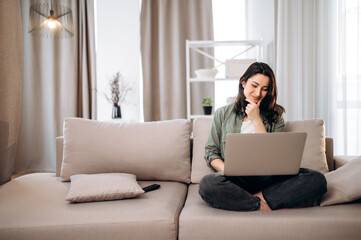 A young gorgeous girl in casual wear sits on a couch and spends time in network chatting with friends and shopping online  using a laptop