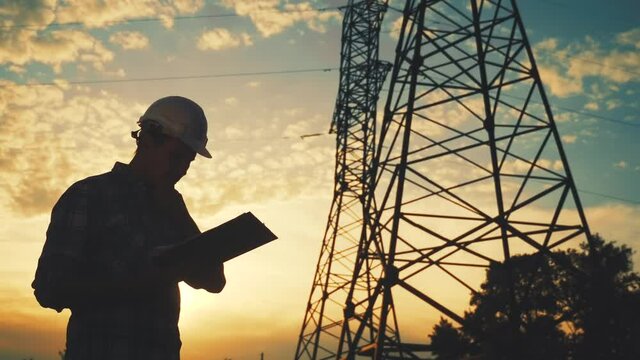 electrical engineer worker in helmet silhouette working at sunset near the tower with electricity. business energy technology industry concept. electrician studying reading documents on a tablet