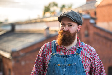 Portrait of brutal bearded stylish craftsman wearing blue overalls, checked shirt and cap in...