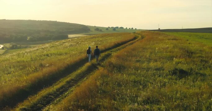 Expanses of meadows and fields, two tourists walk with backpacks on foot. Aerial filming. Beautiful countryside landscape, travelers are walking along the road.