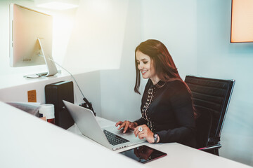 Cheerful beautiful woman receptionist with a long hair is smiling while sitting on armchair surrounded by gadgets and using a laptop indoors of a bright lobby of clinic or business office, or hotel