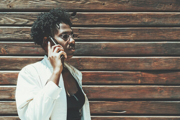 An elegant young black woman in a fancy sunglasses and a white trench is talking on the phone while standing in front of a wooden wall with timber stripes; a copy space place on the right for a text