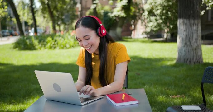 Happy college asian student girl with earphones, backpack, red notebook using laptop and smiling at campus cafe.