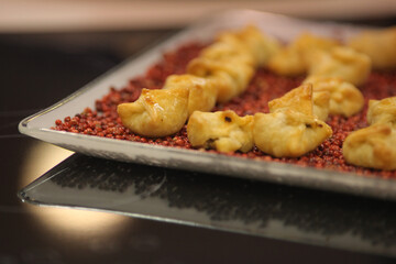 Aperitivo feito com massa folhada e recheio de queijo cremoso, servido numa bandeja sobre cama de pimenta rosa.