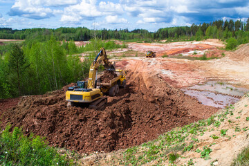 Clay quarry. The yellow excavator digs red clay with a large bucket and puts it into the truck.