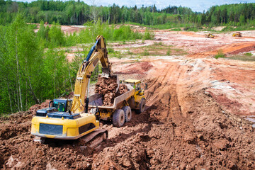 Quarry with red clay. Big yellow excavator extracts clay with stacks in a truck. With place for...