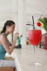 Focus on a glass of cocktail with woman drinking cocktail in the background
