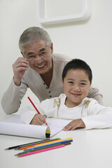 Boy smiling while drawing, senior man standing behind him