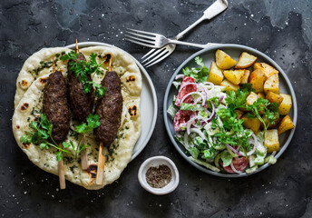 Meat kebab skewers, garlic tortillas naan, yogurt dressing vegetable salad, roasted potatoes - delicious lunch, tapas in the mediterranean style on a dark background, top view
