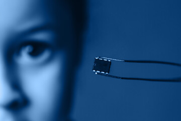 A pair of tweezers holds a identification microchip against the background of the face. The concept...