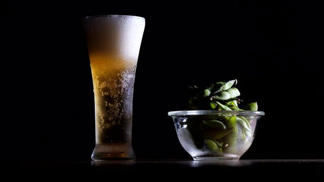 Glass of beer and edamame. Pour beer and foam. Black background. Alcohol and green soybeans. Horizontal angle. Cool