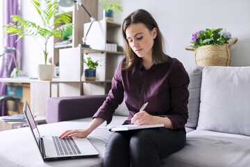 Young woman working remotely at home, sitting on sofa with laptop