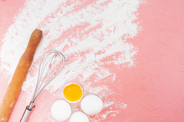 white eggs, a wooden rolling pin, a metal whisk and scattered flour on a delicate pink background. kitchen utensils for baking, dough preparation