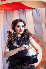 A brunette in a circus doll costume in a black dress takes out a white rabbit from a cylinder. Assistant magician, illusionist, a beautiful girl with art make-up, stands in the arena of the tent 