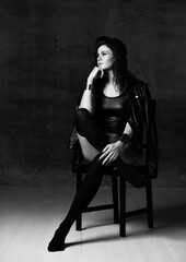 Young thoughtful woman in body underwear, leather jacket, high socks, hat and accessories sits on chair looking at upper corner