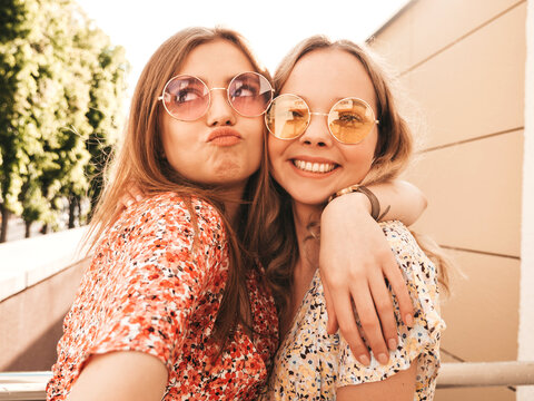 Two young beautiful smiling hipster girls in trendy summer sundress.Sexy carefree women posing on the street background in sunglasses. They taking selfie self portrait photos on smartphone at sunset