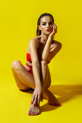Beautiful, young, slim, tanned girl posing in a bright red swimsuit on a yellow background.