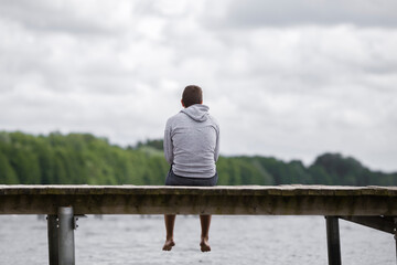 One young adult man sitting alone on edge of footbridge and staring at lake, green trees and cloudy...