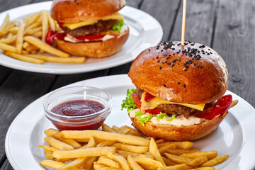 Two hamburgers with beef cutlet, cheese, french fries, tomato, salad, sauce and ketchup on a white plate and a dark wooden background