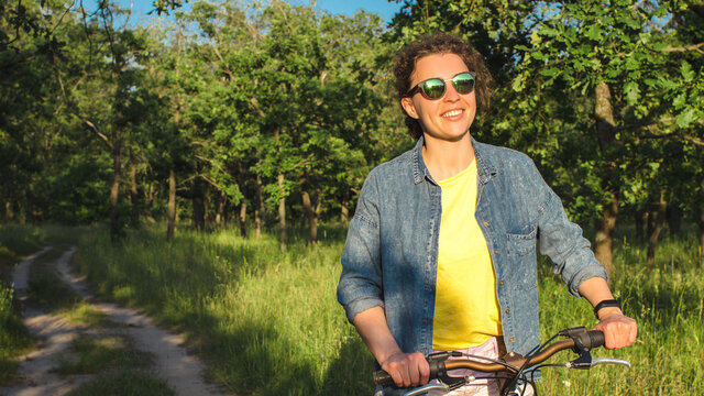 Beautiful smiling young woman riding bicycle in park countryside.Happy brunette girl in sunglasses,summer clothes, enjoying day on bike on green trees background outdoor.Sport,health lifestyle,relax