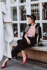 Stylish young woman blonde in a hussar jacket, black hat, jeans, bananas and red shoes in the city near the window of the restaurant. Modern fashion. Soft selective focus.