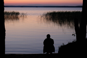  Sunset lake blurry silhouette of a young man looking at the lake, beautiful seascape with sunset. Outdoor summer lake view with dark silhouette. Man looks at the sunset lake. Scenic landscape.
