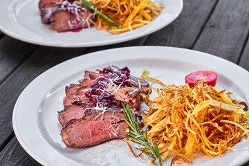 Sliced baked beef with french fries, sauce, tomatoes and rosemary on a white plate and a dark wooden background