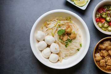 Rice stick noodles with fish ball in white bowl. Garlic cracklings with lard, lime and coriander with spring onion various ingredients and condiment on table.