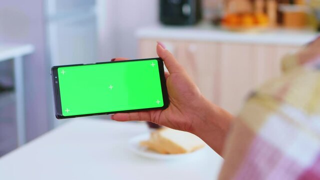 Woman watching a video on phone with green screen in kitchen during morning. Green screen chroma mock up isolated mockup background ready to be replaced with your text, logo or advertisement. Using