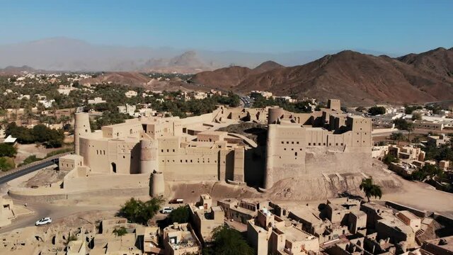 Bahla fort in Oman, famous touristic landmark surrounded by an arid landscape composed of sands, palms, medieval architecture, mountain ranges, under a blue sky