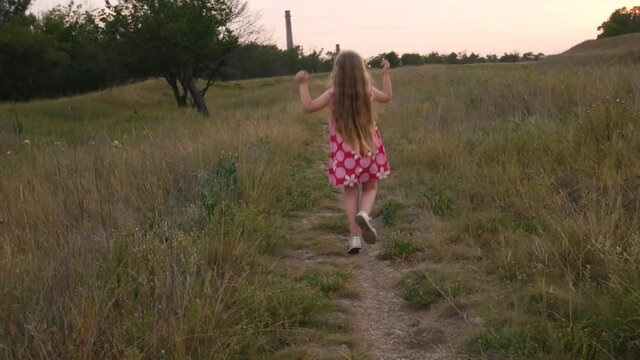 Adorable little girl in pink summer dress walking and jumping in grass and flowers field. Happy child with long blonde wavy hair on green countryside landscape hiking. Local travel concept 4K.