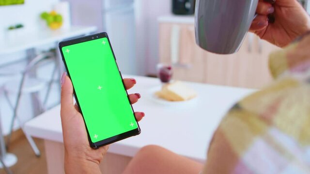 Woman using phone with green screen and drinking coffee in kitchen. French pastry freshly baked on table top. Green screen chroma mock up isolated mockup background ready to be replaced with your text