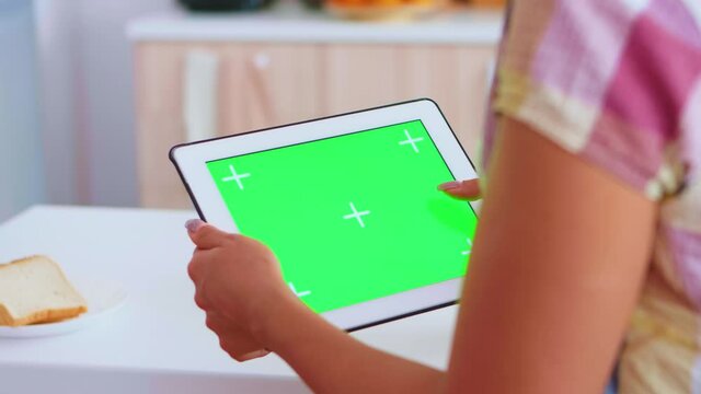 Woman browsing on tablet computer with green screen in kitchen. Green screen chroma mock up isolated mockup background ready to be replaced with your text, logo or advertisement. Using internet