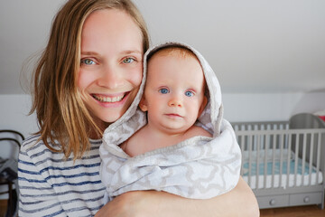 Happy mom holding cute baby wrapped in hooded towel after bathing. Front view. Child care or bathing concept