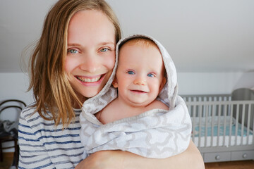 Cheerful mom holding happy baby wearing hooded towel after bathing or shower. Front view. Child care or bathing concept