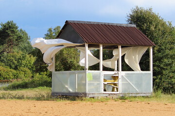 Obraz premium Empty abandoned gazebo with white curtains fluttering in the wind on a deserted Park sand beach against the background of a green trees on a Sunny summer day, windy stormy weather