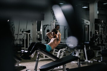 Fototapeta na wymiar Handsome bearded young man with muscular wiry naked torso doing abs exercises during sport workout training in modern dark gym. Concept of healthy lifestyle.