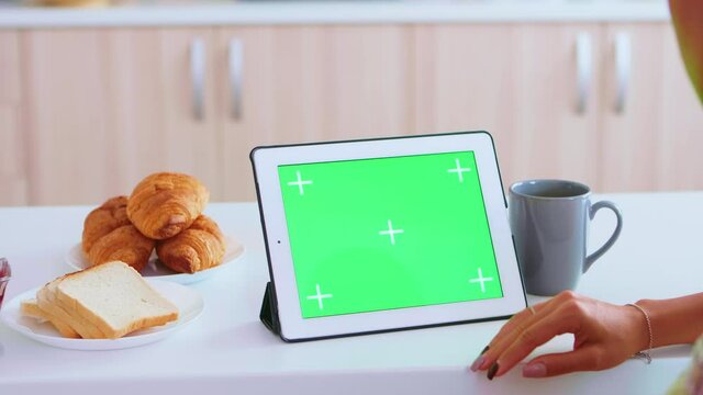 Woman zooming in on tablet pc with green screen during breakfast in kitchen. Tasty french croissants. Green screen chroma mock up isolated mockup background ready to be replaced with your text, logo