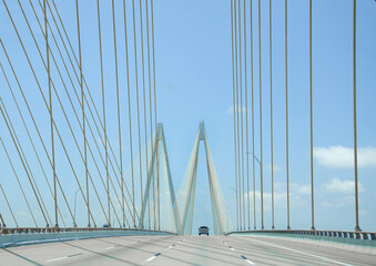 Driving over large cable suspension bridge, with a clear view of the cables and supports and M shaped structure. There is a blue sky and very little traffic. 