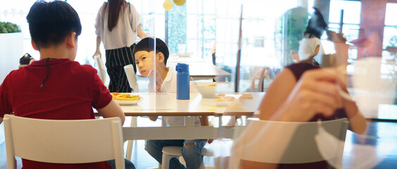Cute little Asian boy talk to his brother who sit on other side of table in food court with clear divider / barrier on table. New normal, Social & physical distancing during Covid-19 pandemic
