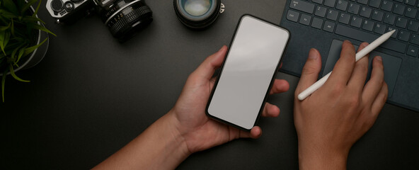 Male holding mock-up smartphone and working on digital tablet on dark office desk