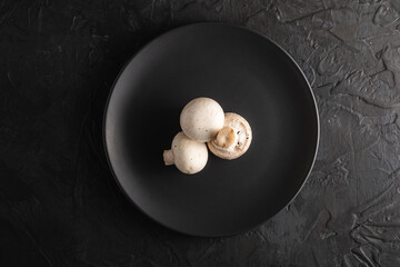 Three champignon mushrooms healthy food on black plate on dark black textured background, top view
