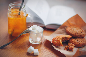 There is a small breakfast on the wooden table - sea buckthorn morse in a glass mug, sugar cubes in a cup and oatmeal cookies. And next to it is an open book. ©  Valeri Vatel