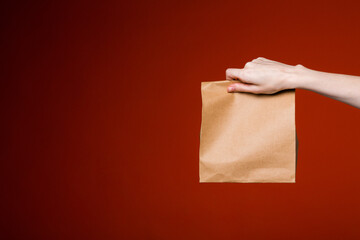 Elongated Female hand holds a paper bag on a red background. Food and gift delivery concept.