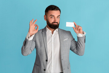 Confident corporate employee holding empty business card and showing ok gesture on blue background, space for design