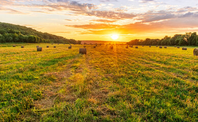 Fototapeta premium Scenic view at beautiful sunset in a green shiny field with hay stacks, bright cloudy sky , trees and golden sun rays with glow, summer valley landscape